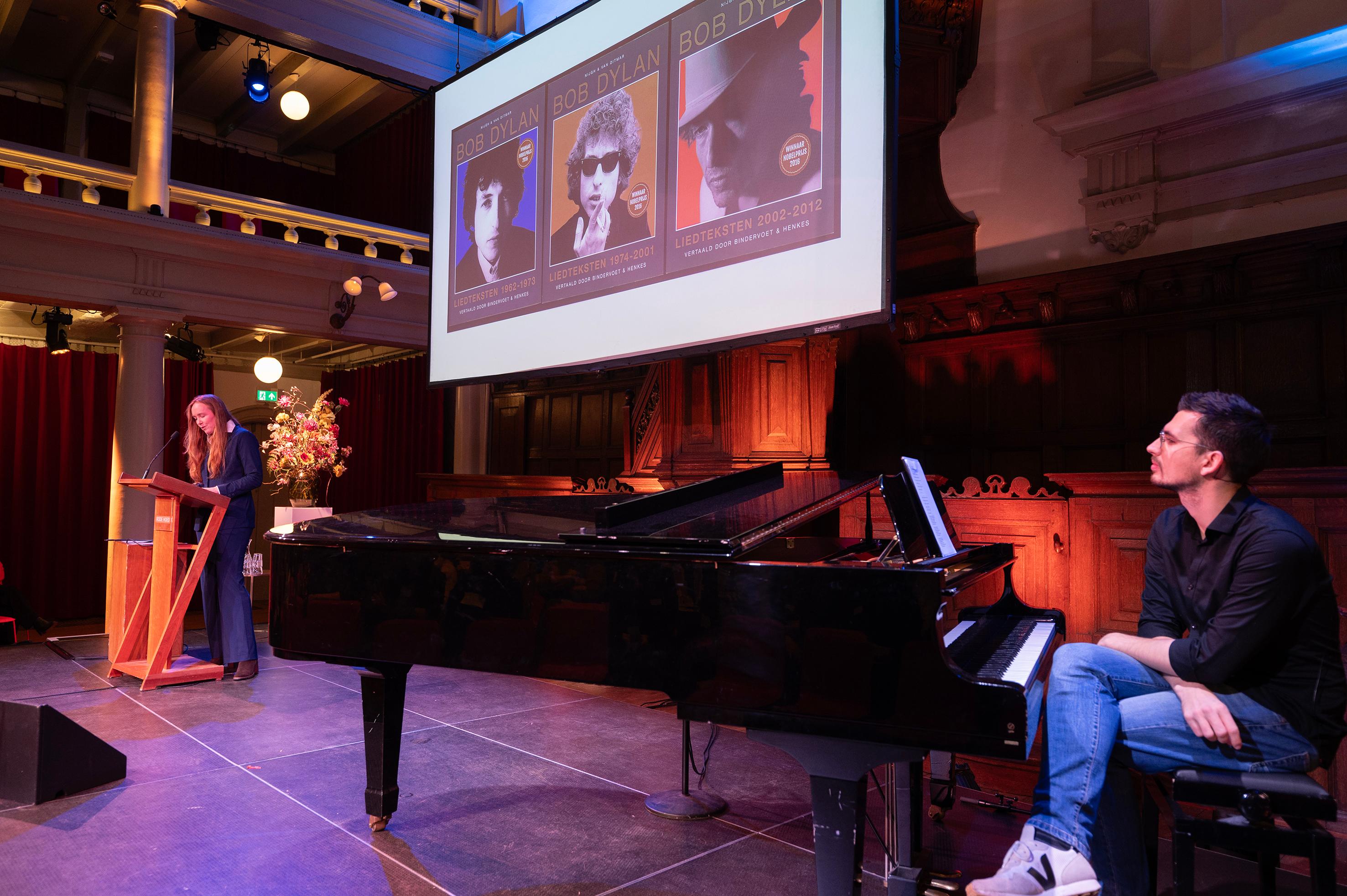 Vrouw staat achter een spreekgestalte en man achter een vleugel, op een podium